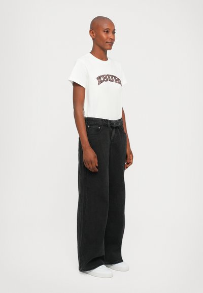 Camiseta blanca de algodón con un gráfico impreso y jeans de mezclilla negros de pierna ancha. El conjunto se complementa con zapatillas deportivas blancas.