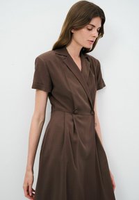Robe marron à manches courtes avec un col plongeant, double boutons, jupe plissée et texture de tissu lisse.