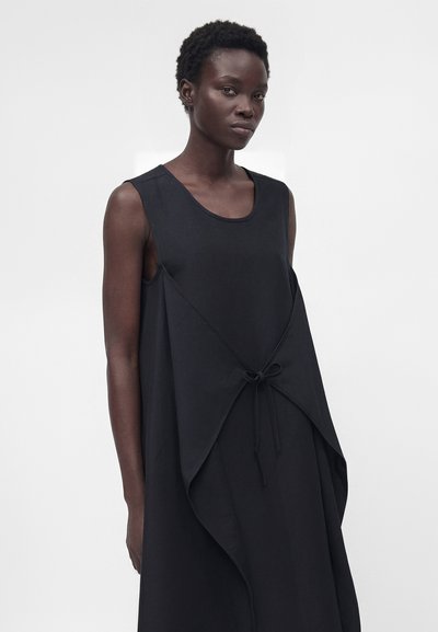 Γυναίκα με κοντά σγουρά μαλλιά που φοράει μαύρο αμάνικο φόρεμα με ασύμμετρη επικάλυψη δεμένη σε φιόγκο στη μέση, στέκεται μπροστά σε απλό φόντο.
