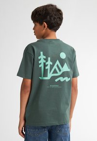Groene katoenen t-shirt met een mintgroene graphic van bomen, bergen en een zon op de achterkant, met tekst onderaan. Casual fit en korte mouwen.
