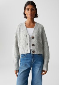Donna che indossa un cardigan a maglia spessa grigio chiaro con grandi bottoni, una maglietta bianca e jeans blu a vita alta su uno sfondo semplice.
