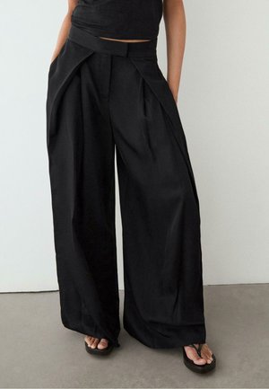 Personne portant un pantalon noir taille haute à jambes larges avec des plis sur le devant et des tongs noires, debout sur un sol gris contre un mur blanc.