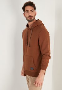 Sudadera con capucha marrón, con un bolsillo frontal, capucha con cordón y puños de canalé. Presenta una etiqueta en el dobladillo inferior. Textura suave, diseño casual.