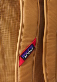 Close-up of tan Patagonia backpack straps showing red logo tag with mountain design and woven fabric texture.