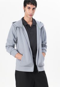 Sudadera gris con cremallera y cordones, de manga larga, con bolsillos frontales y detalles en verde. Llevada sobre una camiseta polo negra. Textura de tela suave.