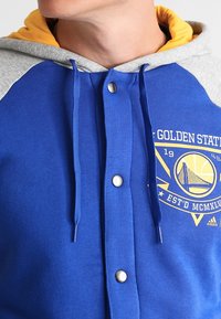 Sudadera azul con mangas grises, que cuenta con una capucha amarilla. El diseño incluye un logotipo gráfico y botones a presión en el frente.