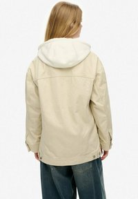 Beige jakke med hvid hætte, der har en løs pasform, sidelommer og knapdetaljer. Stoffet virker glat med en subtil struktur.