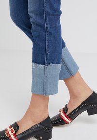 Denim cropped jeans with frayed hems and light blue cuffs paired with black shoes featuring a red and white striped accent and studded detail.