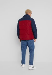 Veste matelassée rouge et marine avec un col montant, un matelassage horizontal et un ourlet élastique, accompagnée de jeans bleus et de baskets blanches.