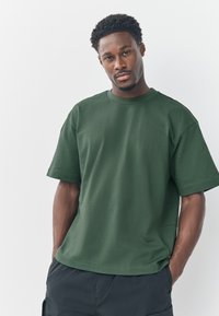 Dunkelgrünes Baumwoll-T-Shirt mit kurzen Ärmeln, rundem Halsauschnitt, lockerem Sitz und minimalen Nähten, kombiniert mit dunklen Shorts.