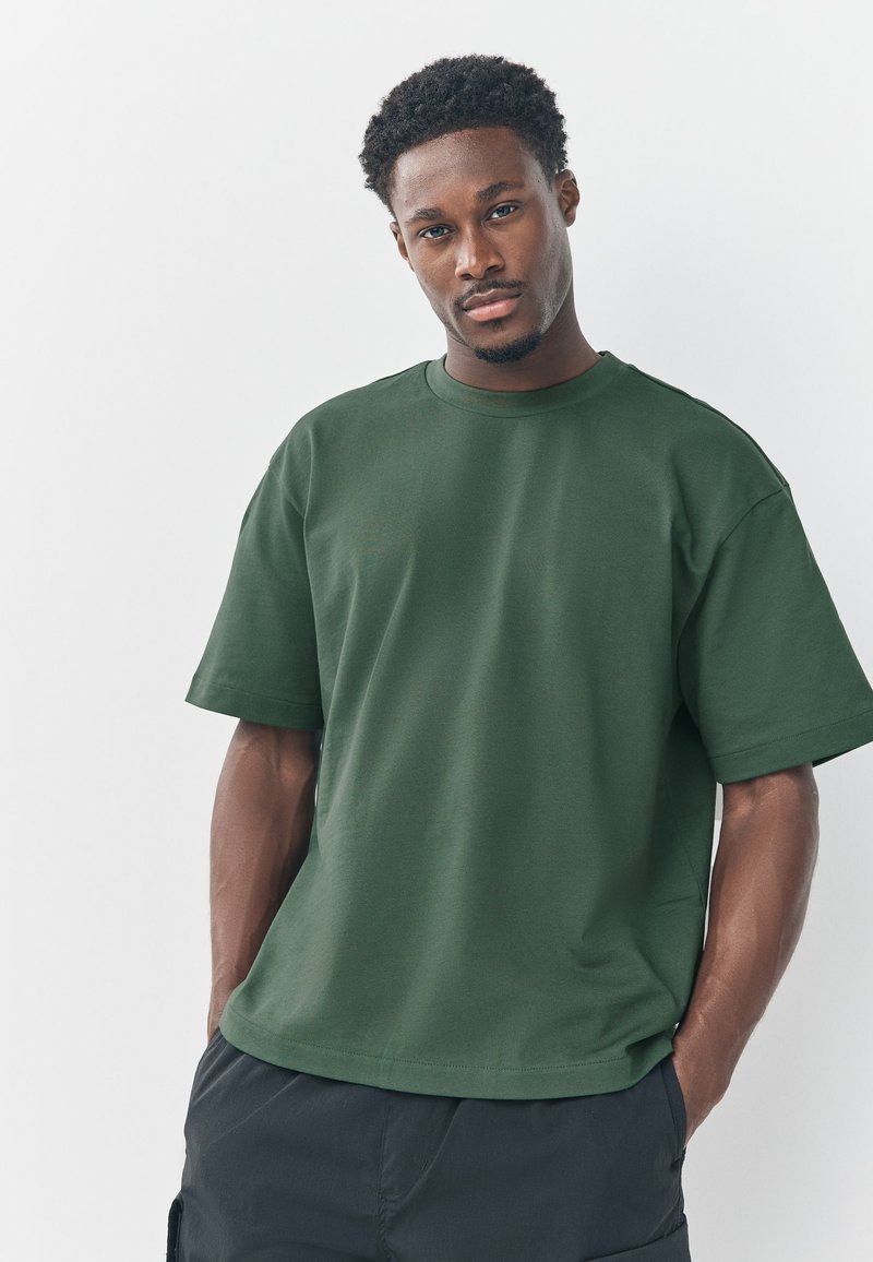 Dunkelgrünes Baumwoll-T-Shirt mit kurzen Ärmeln, rundem Halsauschnitt, lockerem Sitz und minimalen Nähten, kombiniert mit dunklen Shorts.