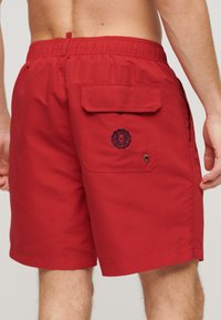 Shorts de bain rouges en tissu léger. Dotés d'une taille élastique, d'une poche arrière avec rabat et d'un logo.