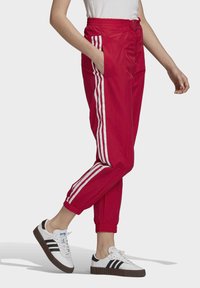 Osoba v červených teplákových kalhotách s bílými postranními pruhy a bílých teniskách s černými pruhy, stojící na jednobarevném pozadí.