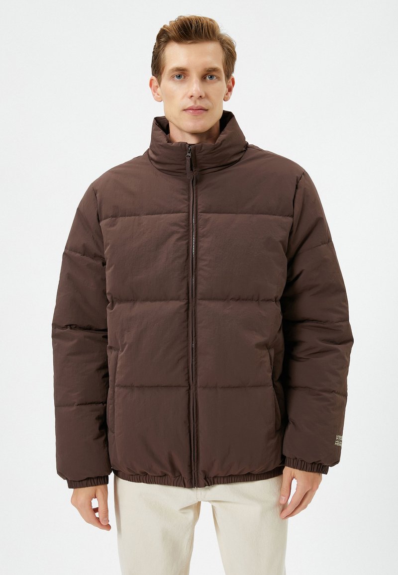Brun pufferjakke med høj krave, lynlås foran og elastiske manchetter. Har quiltet design og slim fit. Stoffet virker glat.