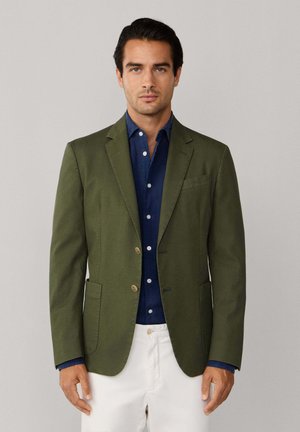 Hombre vistiendo un blazer verde oliva sobre una camisa azul marino abotonada y pantalones blancos, de pie frente a un fondo gris claro liso.