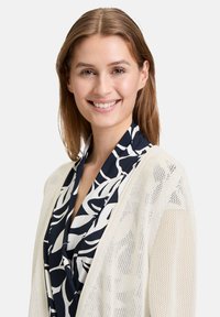 Lange beige cardigan met een gebreide textuur, over een marineblauw en wit blouse met een bloemenpatroon en een gedrapeerde halslijn.