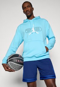 Svetlomodrá mikina s kapucňou a predným vreckom, s čiernym grafickým dizajnom, v kombinácii s námorníckymi šortkami s modrými pruhmi, drží basketbal.