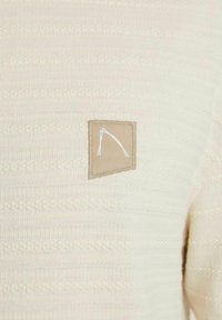 Cremfarbener Strickpullover mit strukturierten Linien und einem rechteckigen beigen Patch mit einem weißen gestickten Design auf der linken Seite.