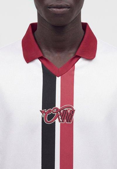 Personne portant une chemise blanche à rayures verticales noires et rouges avec un col rouge, affichant un logo texte stylisé au centre.