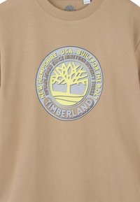 Beige katoenen t-shirt met een ronde, gele en zilveren afbeelding met een boomlogo en tekst over New Hampshire en Timberland.