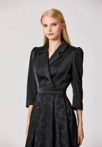 Femme blonde portant un top style blazer en satin noir avec des épaules bouffantes et une jupe à motif floral texturé.
