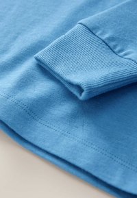 Kék, hosszúujjú ing mandzsetta bordás kötött textúrával és varrott szegéllyel sima anyagfelületen.
