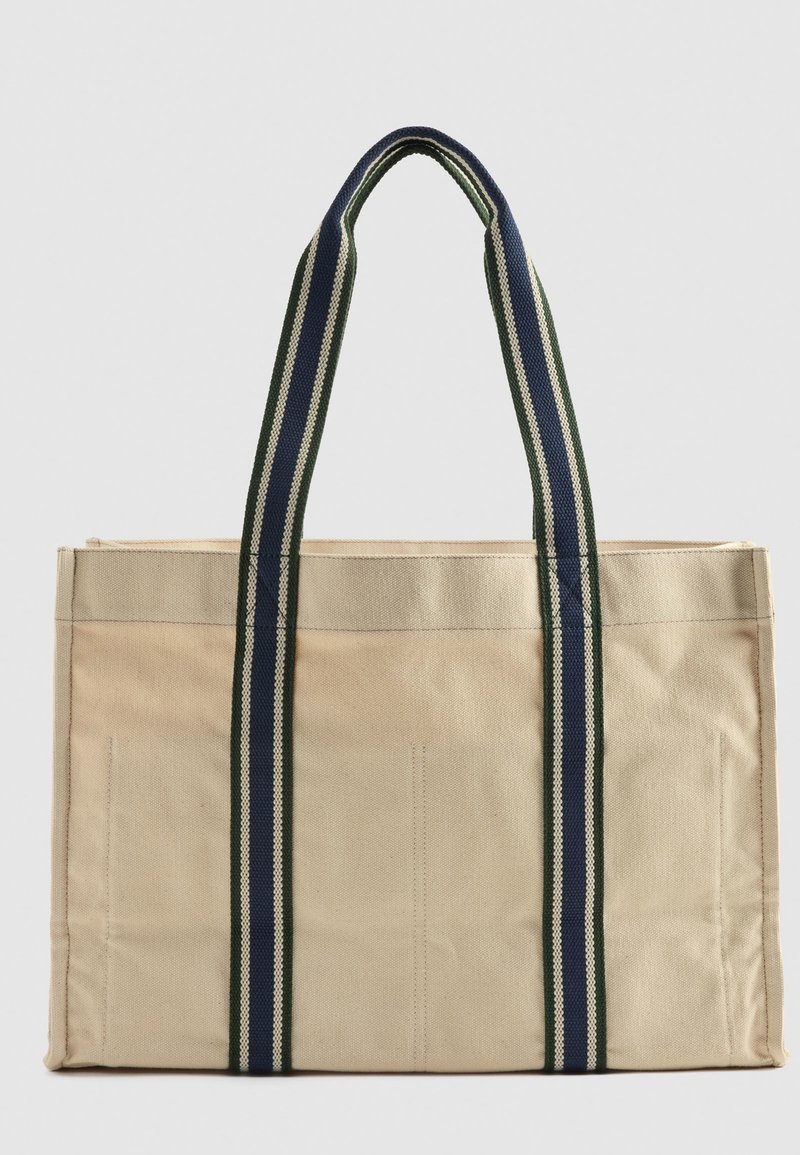 Sac tote en toile beige avec des poignées rayées bleues et vertes, forme structurée et coutures visibles pour le renforcement.
