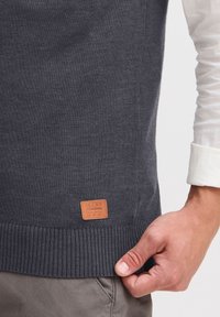 Männerhand passt einen dunkelgrauen Pullover mit einem Lederpatch, auf dem "BLEND" steht, über beigen Hosen und einem sichtbaren weißen Hemdärmel an.