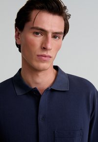 Polo cu un model texturat, de culoare albastru închis, având un guler contrastant, un buzunar pe piept și o deschidere cu nasturi.