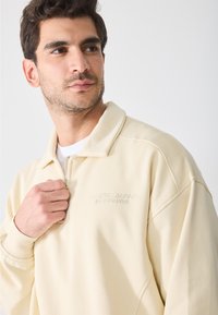 Sudadera de color beige claro con cuello de cremallera, puños de canalé y texto bordado en el pecho. Textura suave y ajuste relajado.