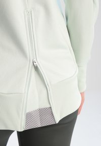 Gros plan d’une veste de couleur claire avec une fermeture éclair latérale partiellement ouverte, révélant une doublure intérieure perforée et un ourlet côtelé sur un pantalon sombre.