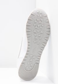 Semelle d'une basket blanche Fila avec des motifs triangulaires et des lignes courbes au niveau du talon, positionnée verticalement contre un coin blanc.