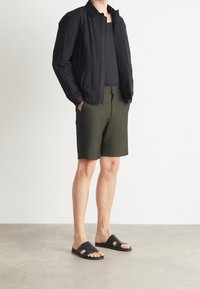 Černá bomber bunda na zip, černý top na ramínka, olivově zelené šortky, černé klouzavé sandály; hladký materiál, minimalistický design, uvolněný střih.