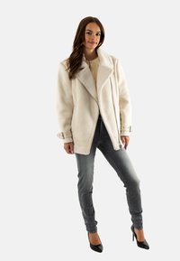 Donna che indossa una giacca in shearling color crema, jeans aderenti grigi e scarpe con tacco nere, in piedi contro uno sfondo chiaro e semplice.