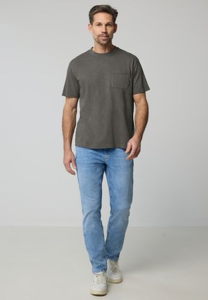 Uomo che cammina in avanti indossando una t-shirt a maniche corte grigio scuro, jeans azzurri e scarpe da ginnastica bianche su uno sfondo neutro.