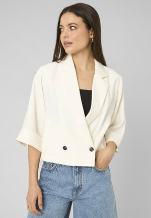 Femme portant un blazer court blanc à manches trois-quarts sur un haut noir et un jean taille haute bleu clair, regardant vers la gauche.