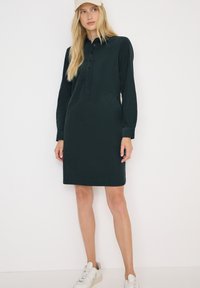Frau in einem dunkelgrünen knöpfbaren Hemdkleid, beigem Hut und weißen Sneakers, die vor einem schlichten weißen Hintergrund steht.