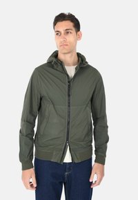 Giacca leggera verde con cappuccio, chiusura a zip frontale, due tasche laterali, polsini elastici e una finitura texturizzata. Indossata sopra una camicia leggera.