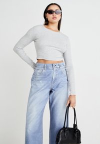 Siv sweatshirt s dolgimi rokavi, svetlo plavi široki jeans in majhna črna torba. Temna sončna očala in gladke teksture tkanin so vidne.