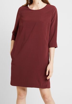 Kobieta ubrana w bordową sukienkę do kolan z rękawami do łokcia i kolczykami z perłami, stojąca na jednolitym tle.