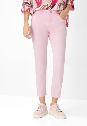 Person iført lyserøde, korte bukser, blomstret bluse og lyserøde sneakers, stående mod en hvid baggrund.