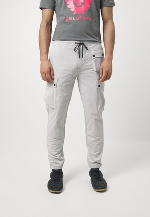 Homme portant un pantalon cargo gris clair avec plusieurs poches et des baskets noires, debout devant un fond blanc uni.