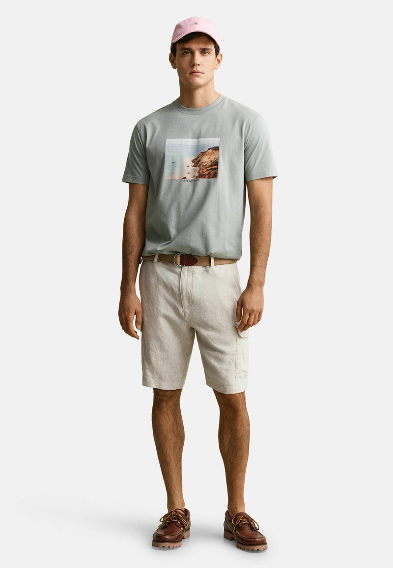 Joven con gorra rosa, camiseta gris con un estampado costero, pantalones cortos beige con cinturón y zapatos náuticos marrones, de pie sobre fondo blanco.