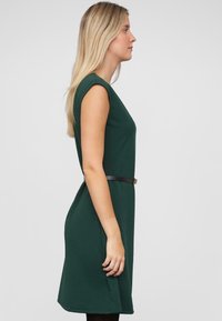 Grünes ärmelloses Kleid mit taillierter Form, betontem Bund durch einen schwarzen Gürtel, glatter Textur und knielangem Saum.