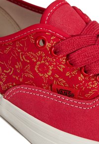Rode canvas sneaker met gouden bloemenpatroon, bovenwerk van gestructureerd suède, ronde rode veteroogjes en witte rubberen zool. Logotag zichtbaar.