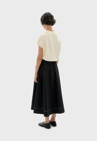 Crème korte mouwen top gecombineerd met een zwarte, lange A-lijn rok met witte stiksels en een zijzak, van achteren weergegeven met zwarte schoenen.