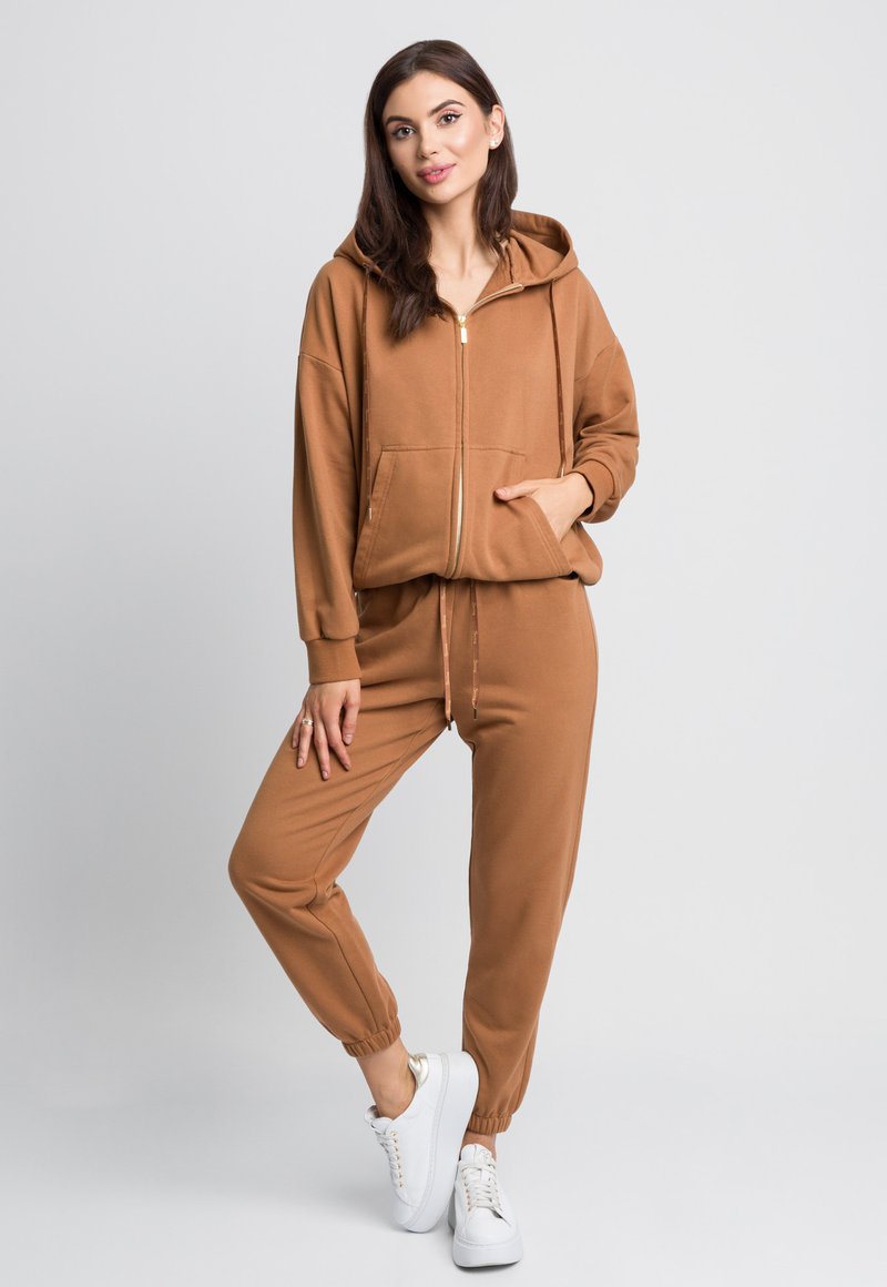 Brązowa bluza rozpinana z kieszeniami z przodu, zestawiona z dopasowanymi spodniami dresowymi z elastycznymi mankietami. Białe sneakersy dopełniają stylizację.