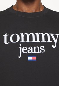 Felpa nera con collo a giro in cotone con testo bianco "tommy jeans" e un piccolo logo rettangolare rosso, bianco e blu sotto il testo.