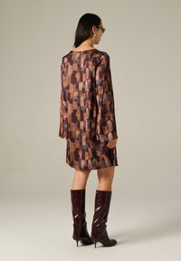 Braunes gemustertes Kleid mit langen Ärmeln, das abstrakte Formen in dunklen Farbtönen zeigt. Das Model trägt knielange burgunderfarbene glänzende Stiefel.