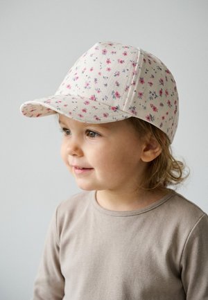 Kleinkind lächelt, trägt eine beigefarbene Kappe mit Blumenmuster und ein hellbraunes Langarmshirt, schaut nach rechts vor hellem einfarbigem Hintergrund.